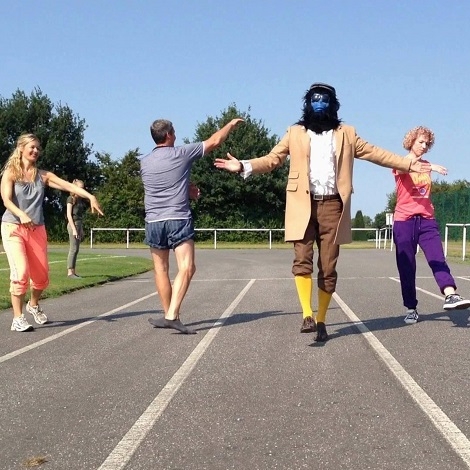 Der blaue Affe Kalle, gekleidet in festlicher Tracht, läuft elegant mit weiteren Personen auf einer Sprintbahn.